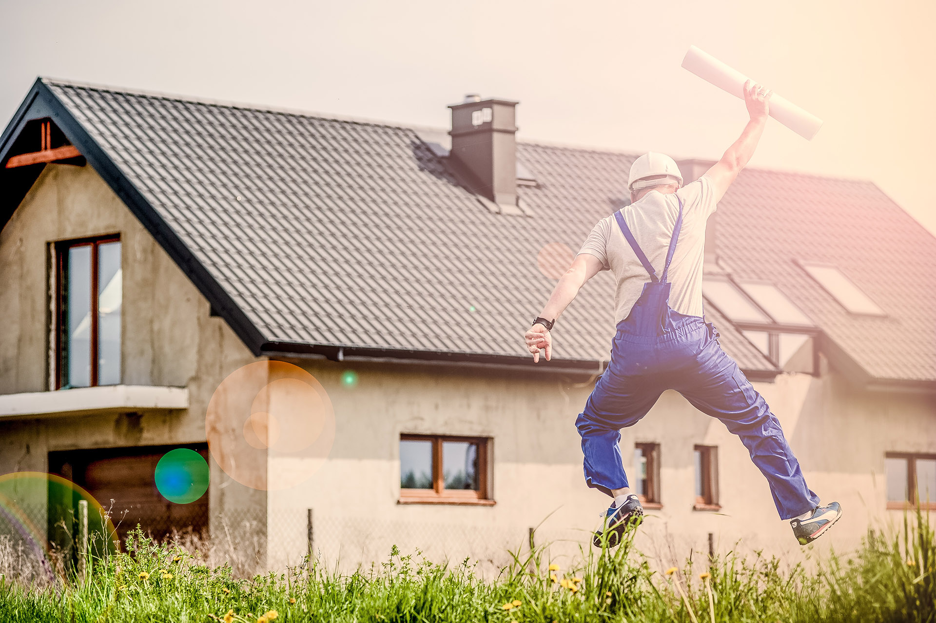 Handwerker springt gut gelaunt mit einer Bauzeichnung vor einem Haus in die Luft